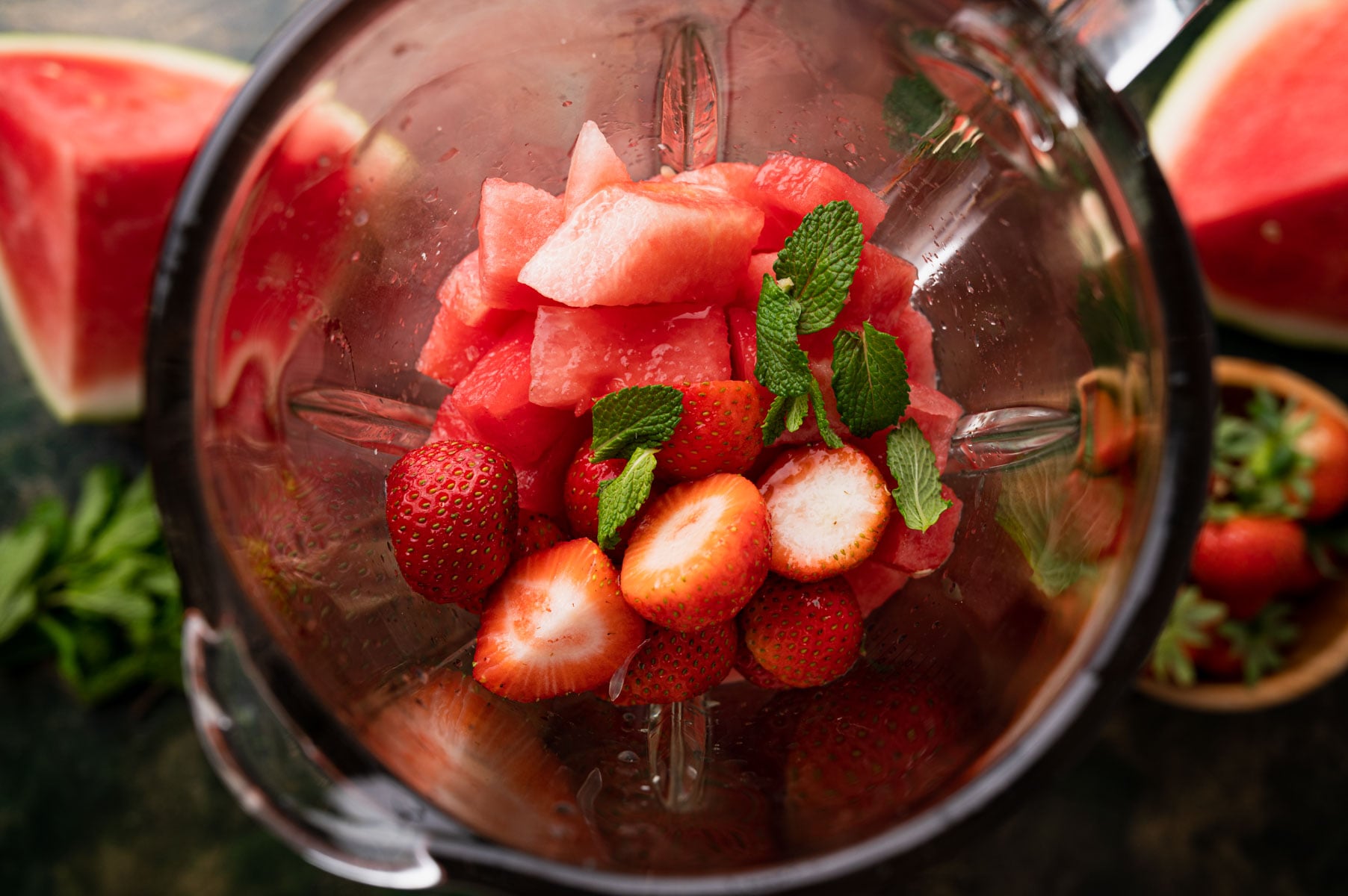 A blender filled with watermelon chunks, strawberries, and mint leaves, ready for blending with more watermelon pieces in the background.