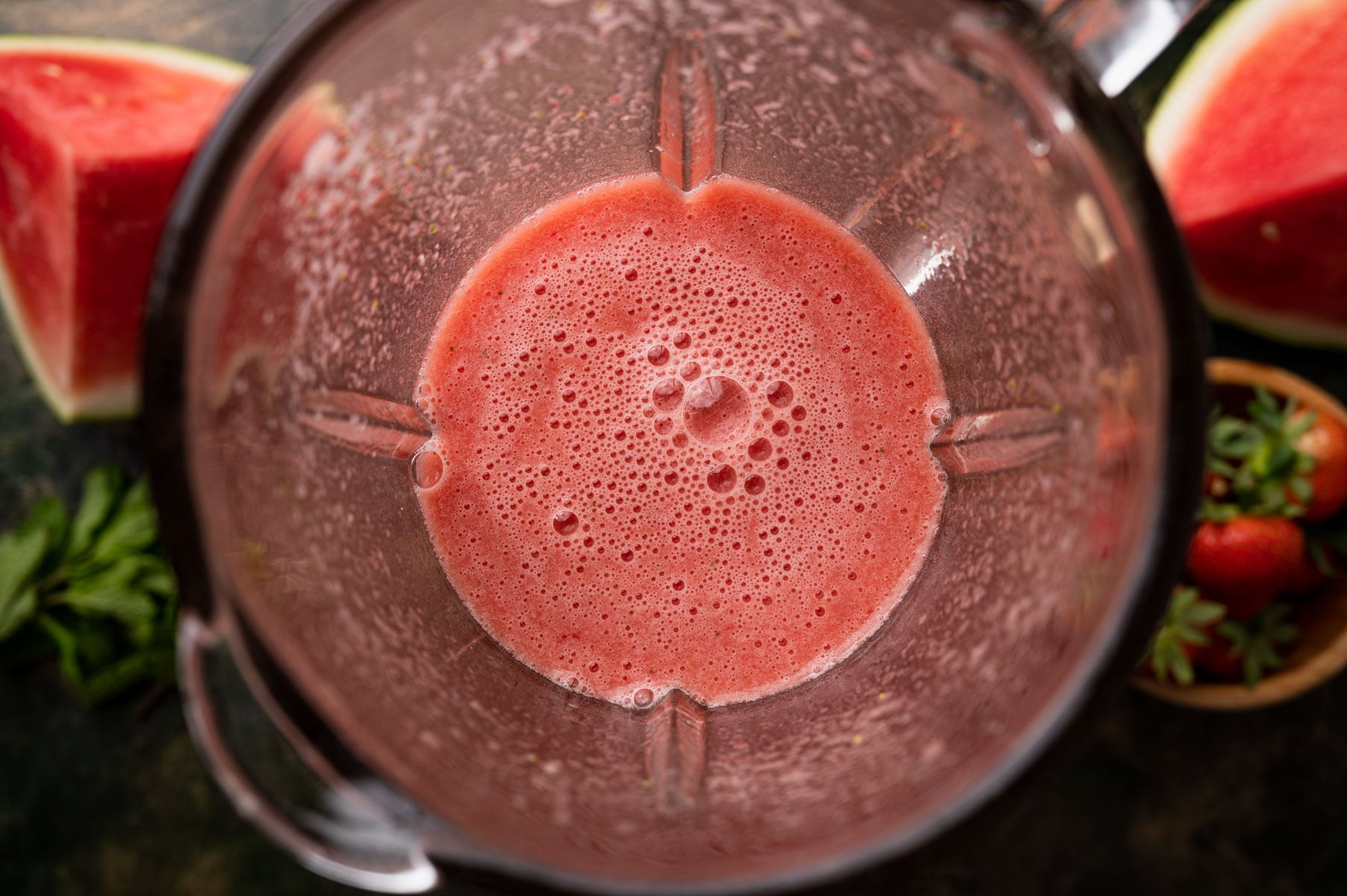 Top view of a blender containing pink, frothy watermelon juice with watermelon slices and strawberries surrounding it.