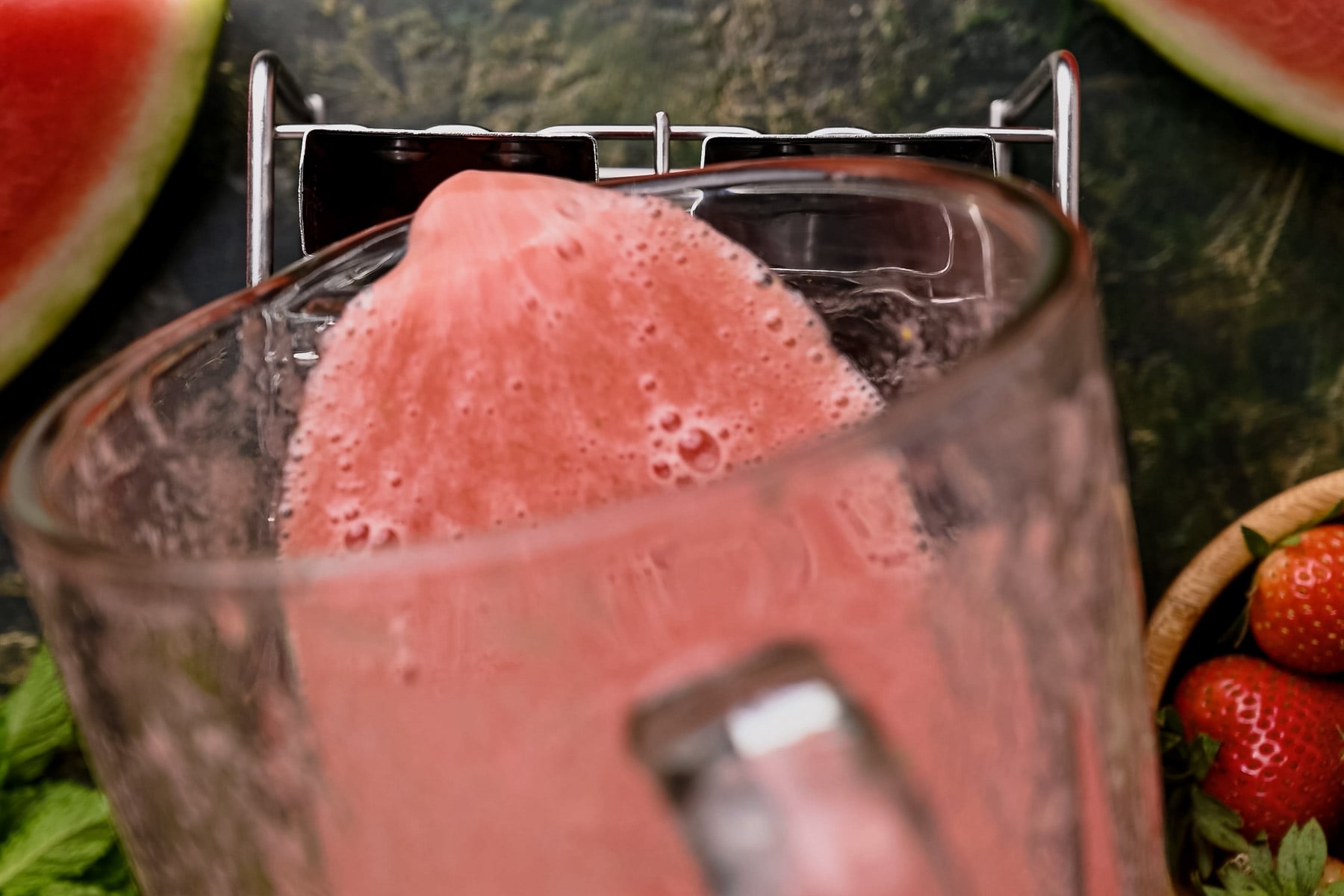 Blender pouring a pink smoothie into a glass with watermelon and strawberries nearby on a green textured surface.