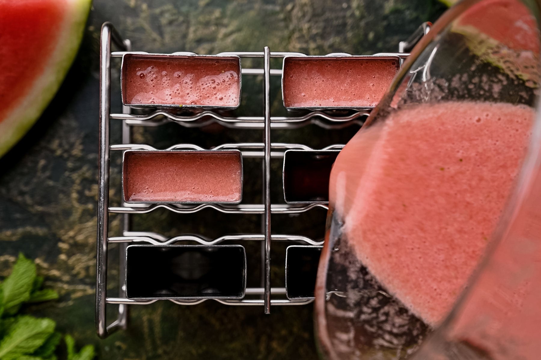 Pouring watermelon smoothie mixture into a metal popsicle mold on a greenish surface.