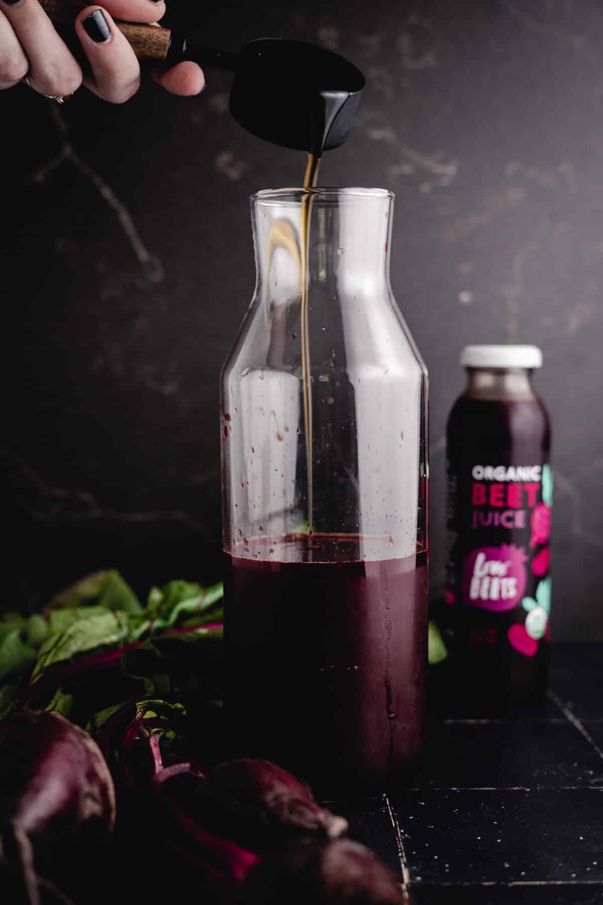 A person pours maple syrup into a clear glass bottle with beet juice inside. A beet juice bottle labeled "Organic Beet Juice" is in the background, along with fresh beets and greens on a dark surface.