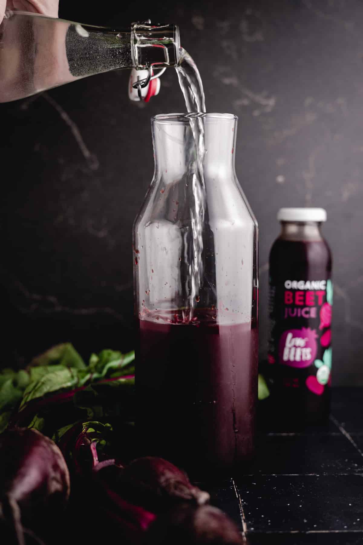 Water is being poured into a glass container containing red liquid. A bottle of organic beet juice stands in the background, and fresh beets with greens are placed nearby.