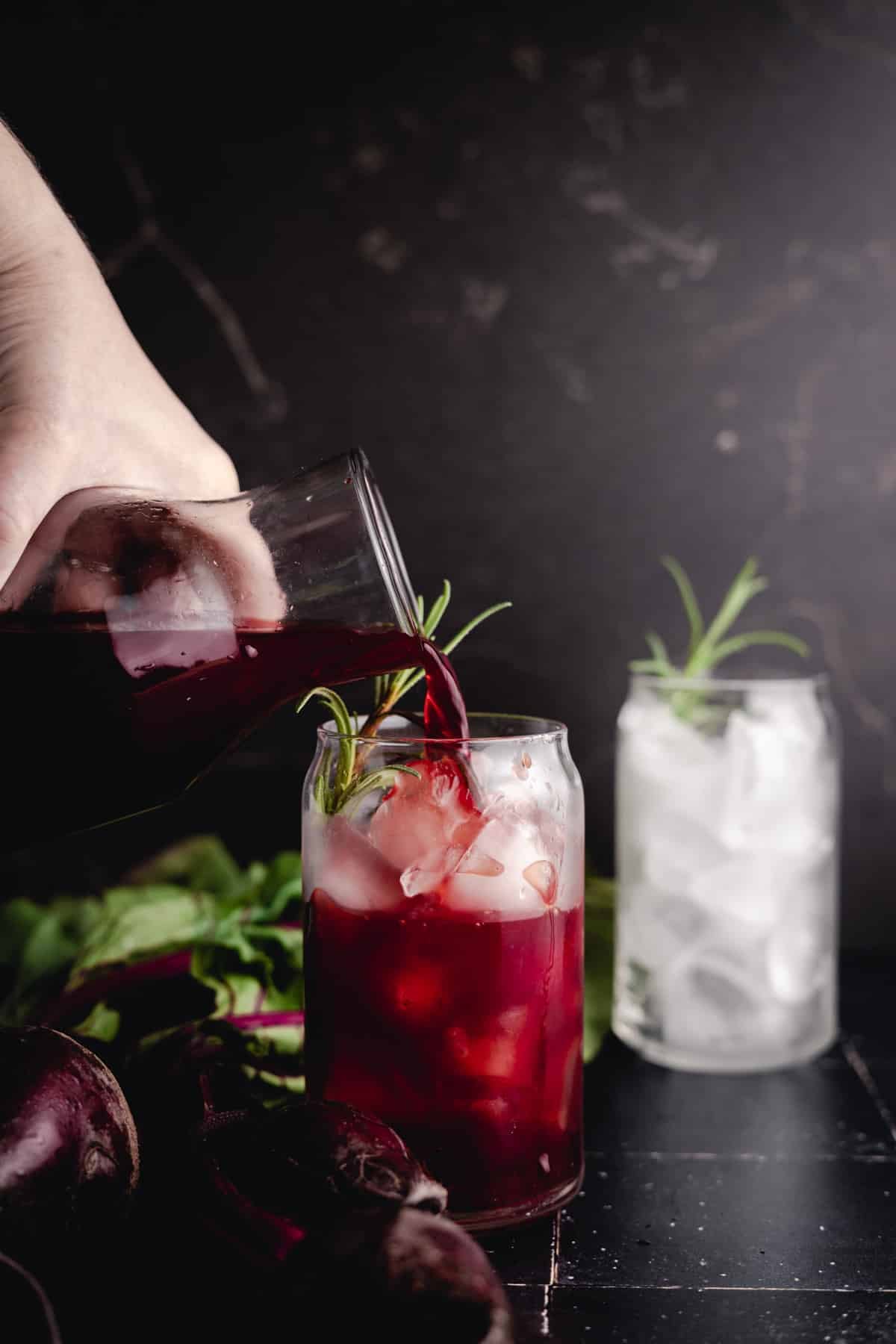 A hand pouring beet lemonade from a glass pitcher into a glass filled with ice, with another glass of ice and a leafy garnish in the background.