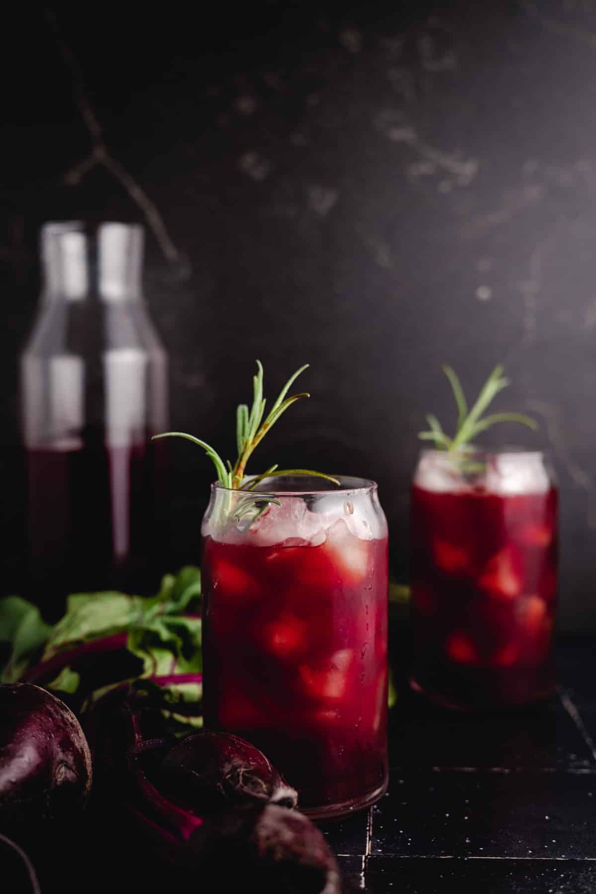 Two glasses of iced beet juice garnished with rosemary sprigs, with a glass pitcher and beetroots in the background.