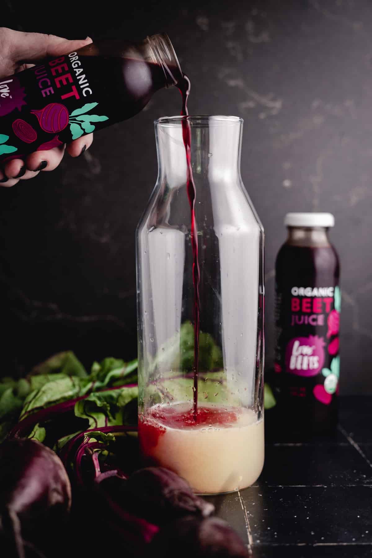 Pouring beet juice from a bottle into a clear glass pitcher, with another bottle and fresh beets placed around it on a dark surface.