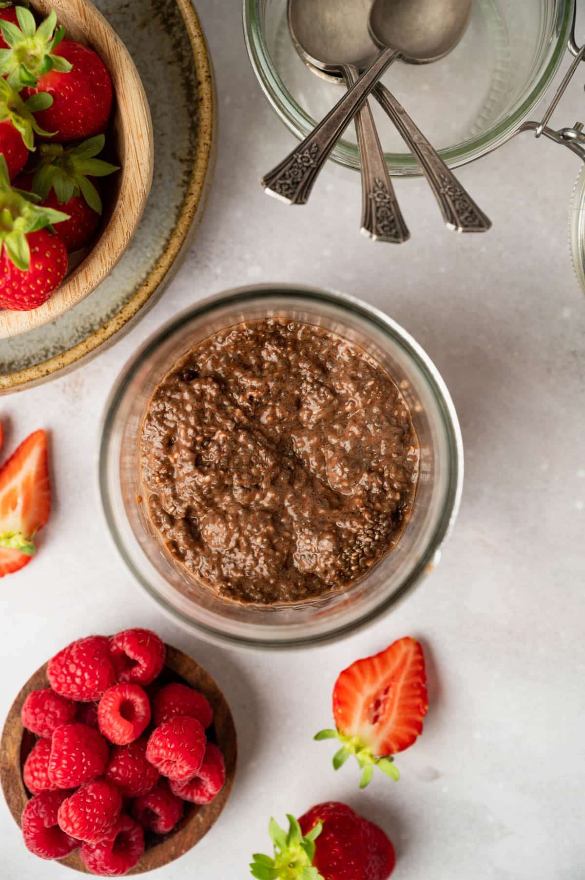 A glass jar of chocolate chia pudding is surrounded by fresh strawberries, raspberries, and silver spoons laid on a light-colored surface.