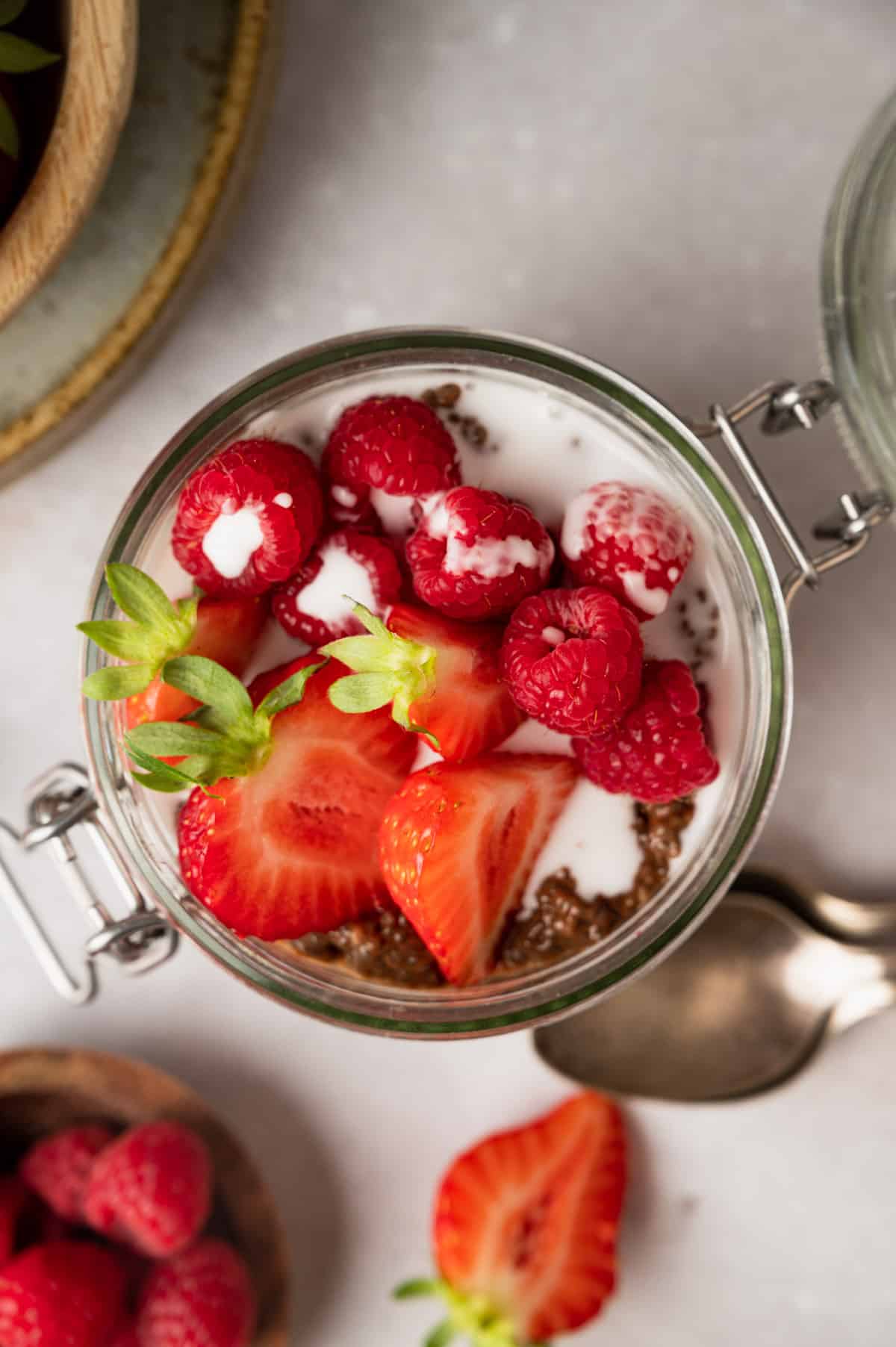 A jar of yogurt topped with chia seeds, fresh strawberries, and raspberries sits on a light surface. A spoon, a bowl of raspberries, and a sliced strawberry are near the jar.