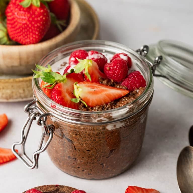 A glass jar filled with chocolate chia pudding topped with strawberries and raspberries, surrounded by sliced strawberries and wooden spoons on a table.