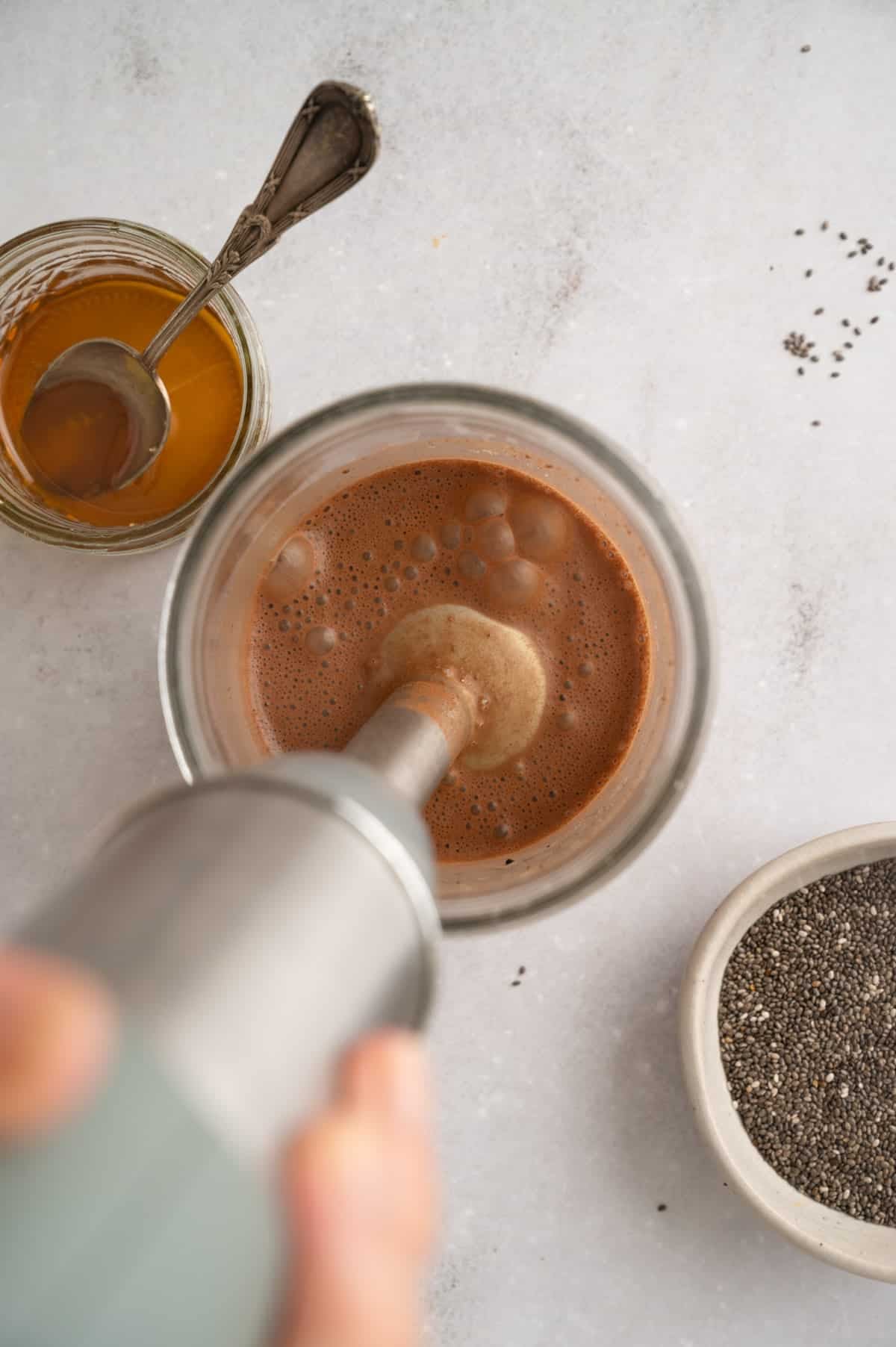 Person blending a brown liquid in a glass with an immersion blender, next to a small bowl of chia seeds and a jar of maple syrup with a spoon.