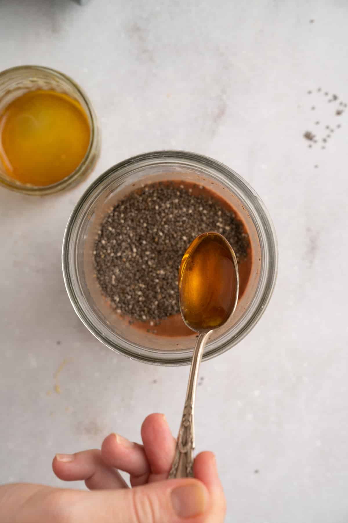 A hand holds a spoon with maple syrup over a jar containing chia seeds and liquid, with another jar containing maple syrup in the background.