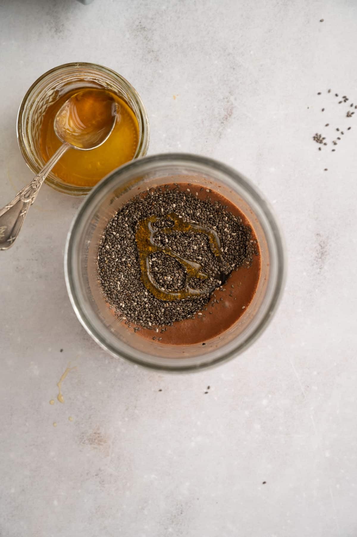 A glass jar containing a mixture of chia seeds and a dark liquid sits next to an empty jar with a spoon coated in a maple syrup on a light-colored surface.