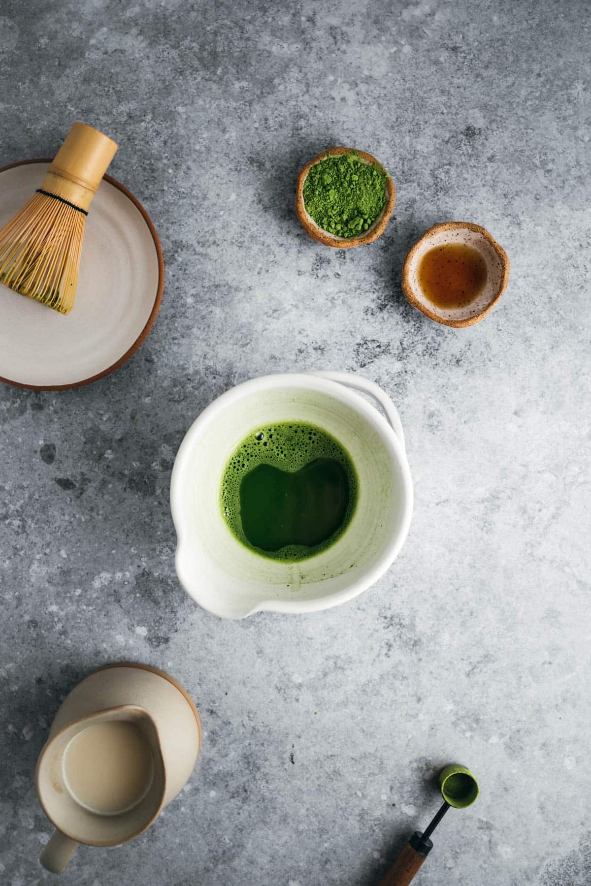 Overhead view of a matcha tea setup on a gray surface. Includes a bowl of whisked matcha, a whisk, matcha powder, a small cup of liquid, a wooden spoon with matcha, and a teapot.