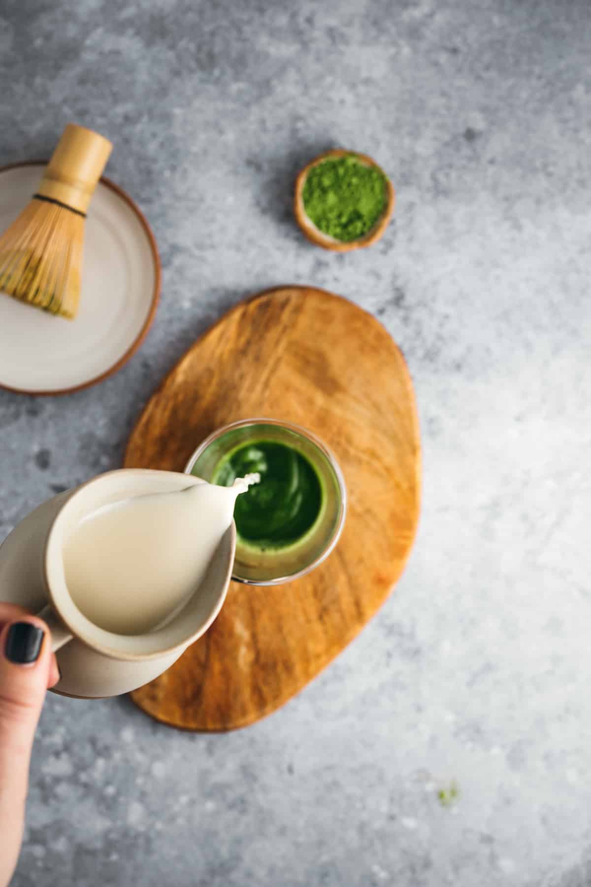 A hand is pouring milk from a small jug into a green matcha beverage on a wooden board, with a matcha whisk and a small bowl of matcha powder nearby.
