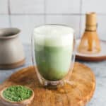 A clear glass mug filled with frothy matcha latte stands on a wooden board. Nearby are a small bowl of matcha powder and a traditional bamboo whisk.