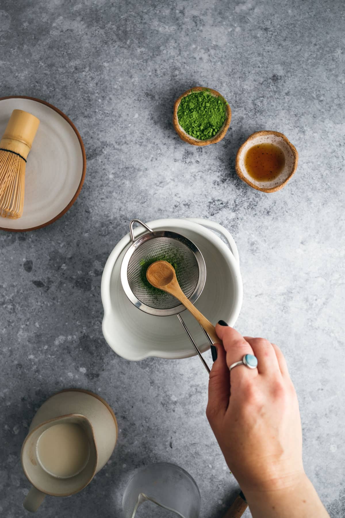 A hand holds a wooden spoon sifting green matcha powder into a white bowl on a gray surface. Nearby are bowls containing more matcha powder, a bamboo whisk, and a small cup of tea.