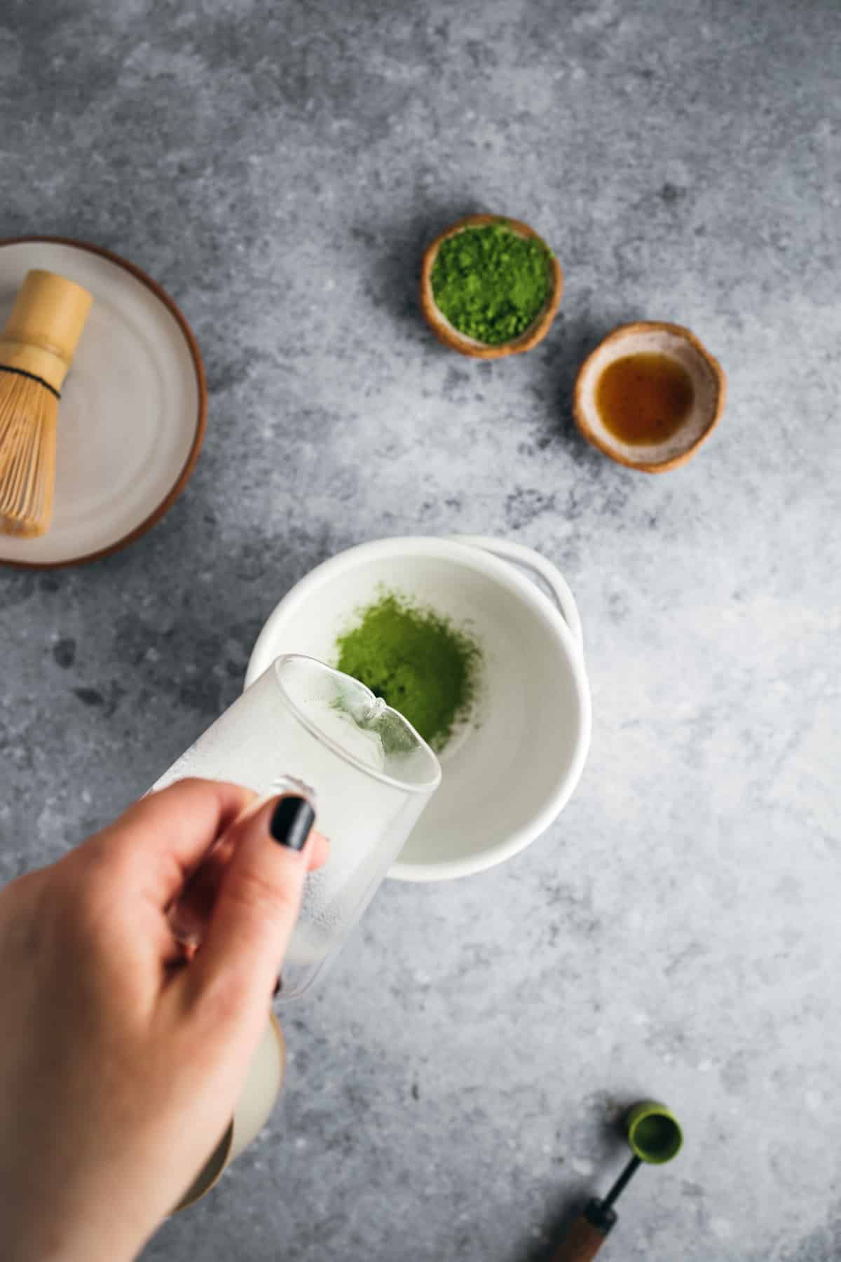 A hand pours liquid into a bowl containing matcha powder. Nearby, two small bowls hold matcha powder and liquid, and a bamboo whisk rests on a saucer. A flat surface serves as the background.
