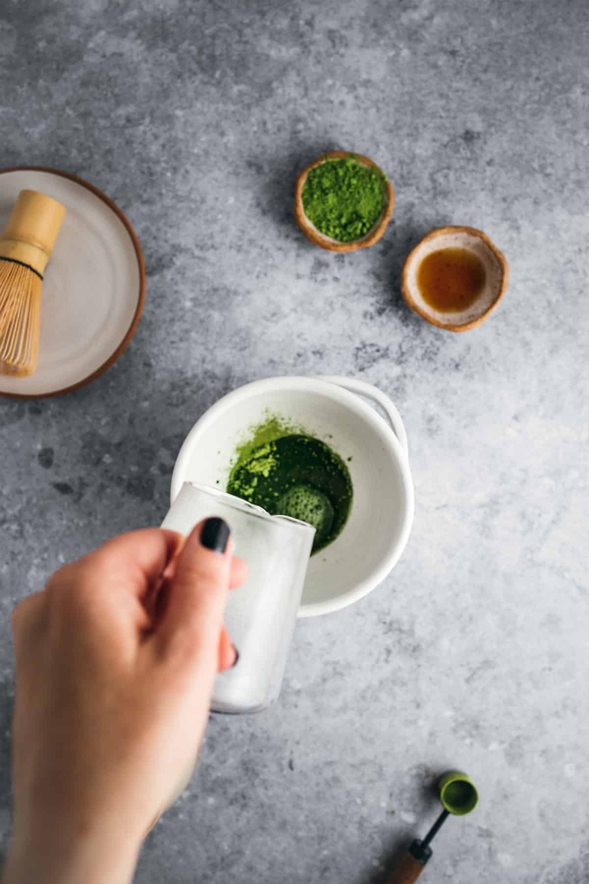 A person’s hand stirring matcha in a white bowl with a whisk, accompanied by bowls of matcha powder and tea on a gray surface.