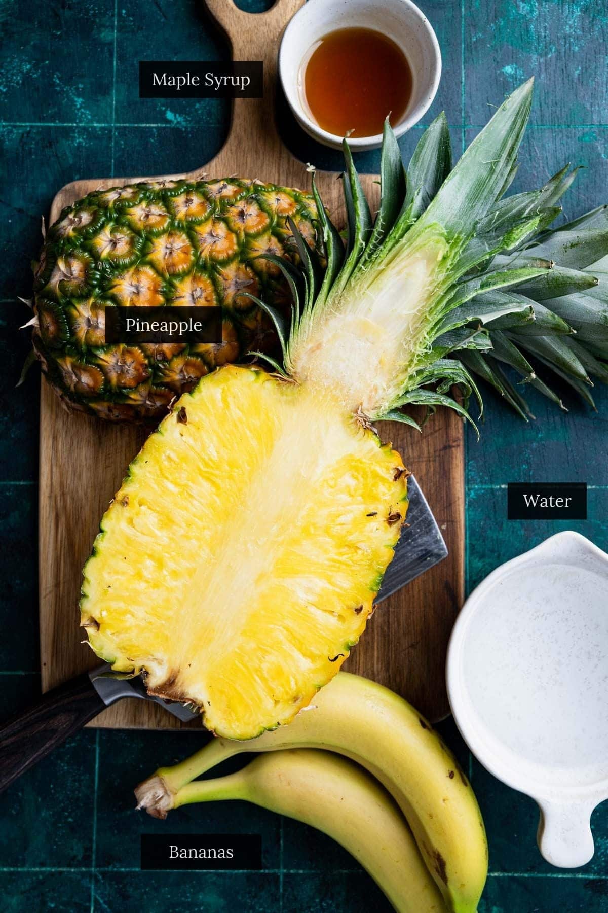 A cutting board with a halved pineapple, bananas, a small bowl of maple syrup, and a pitcher of water—all labeled.