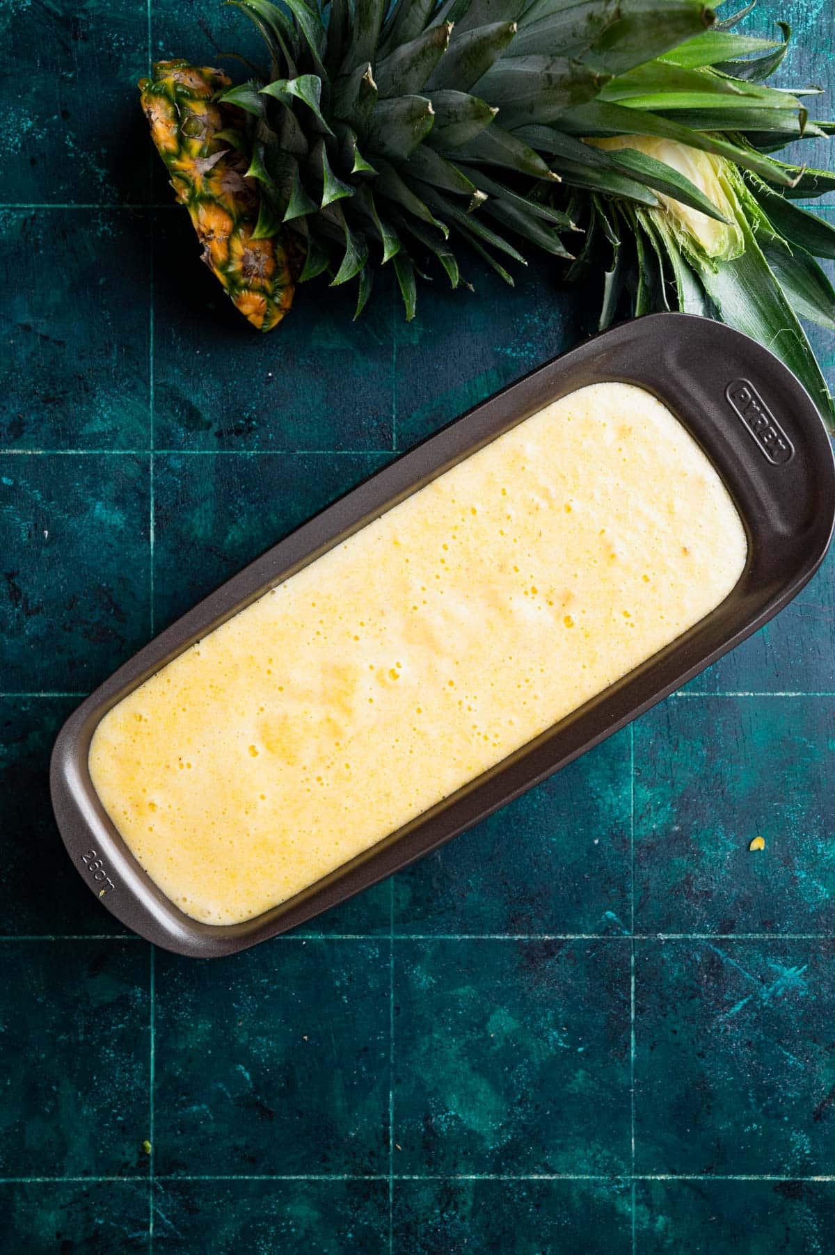 A rectangular baking dish filled with yellow batter on a dark green textured background, with a cut pineapple and its leaves placed above the dish.