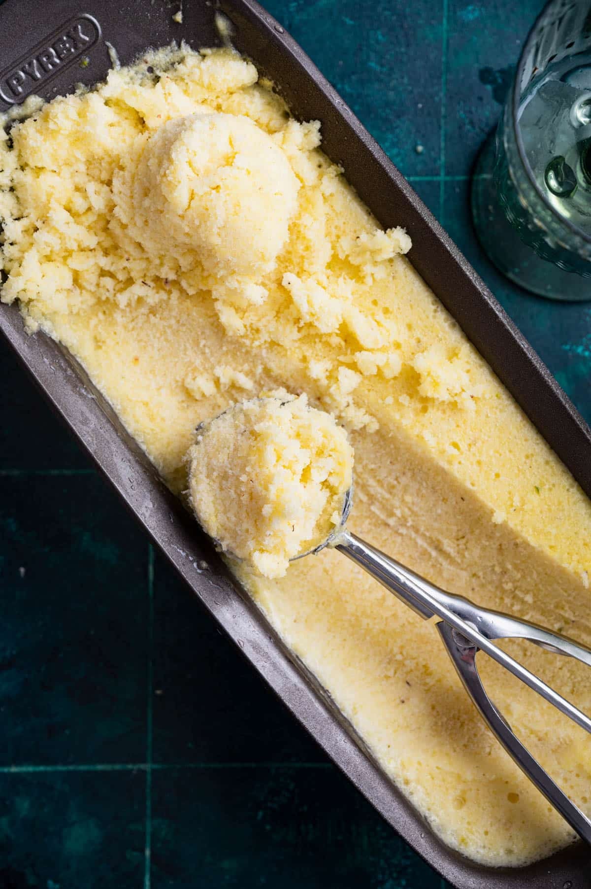 A loaf pan of homemade lemon sorbet with scoops taken out, a metal ice cream scooper resting inside, all set on a green countertop.