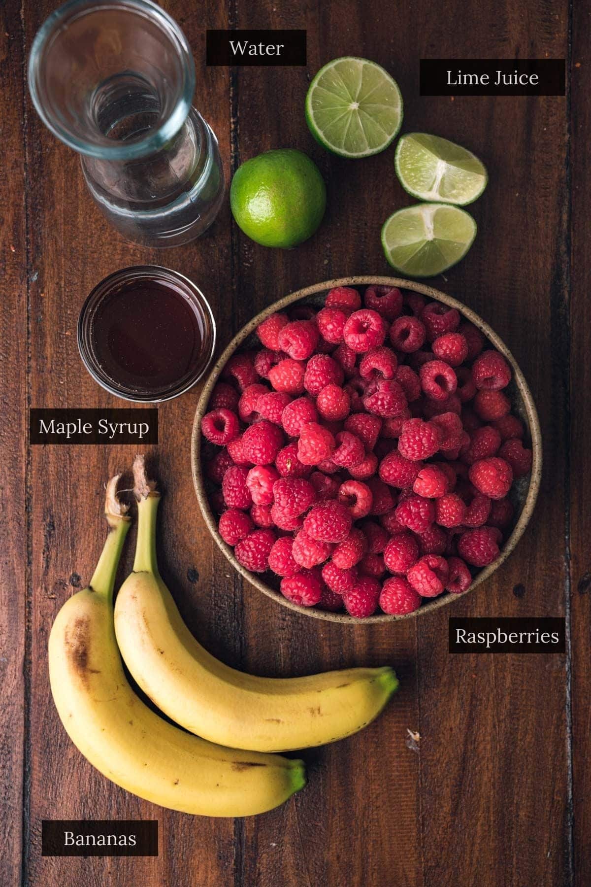 A bowl of raspberries sits on a wooden surface alongside bananas, a jar of maple syrup, a glass of water, and two halved limes labeled as lime juice—a perfect setup for creating refreshing raspberry sorbet.