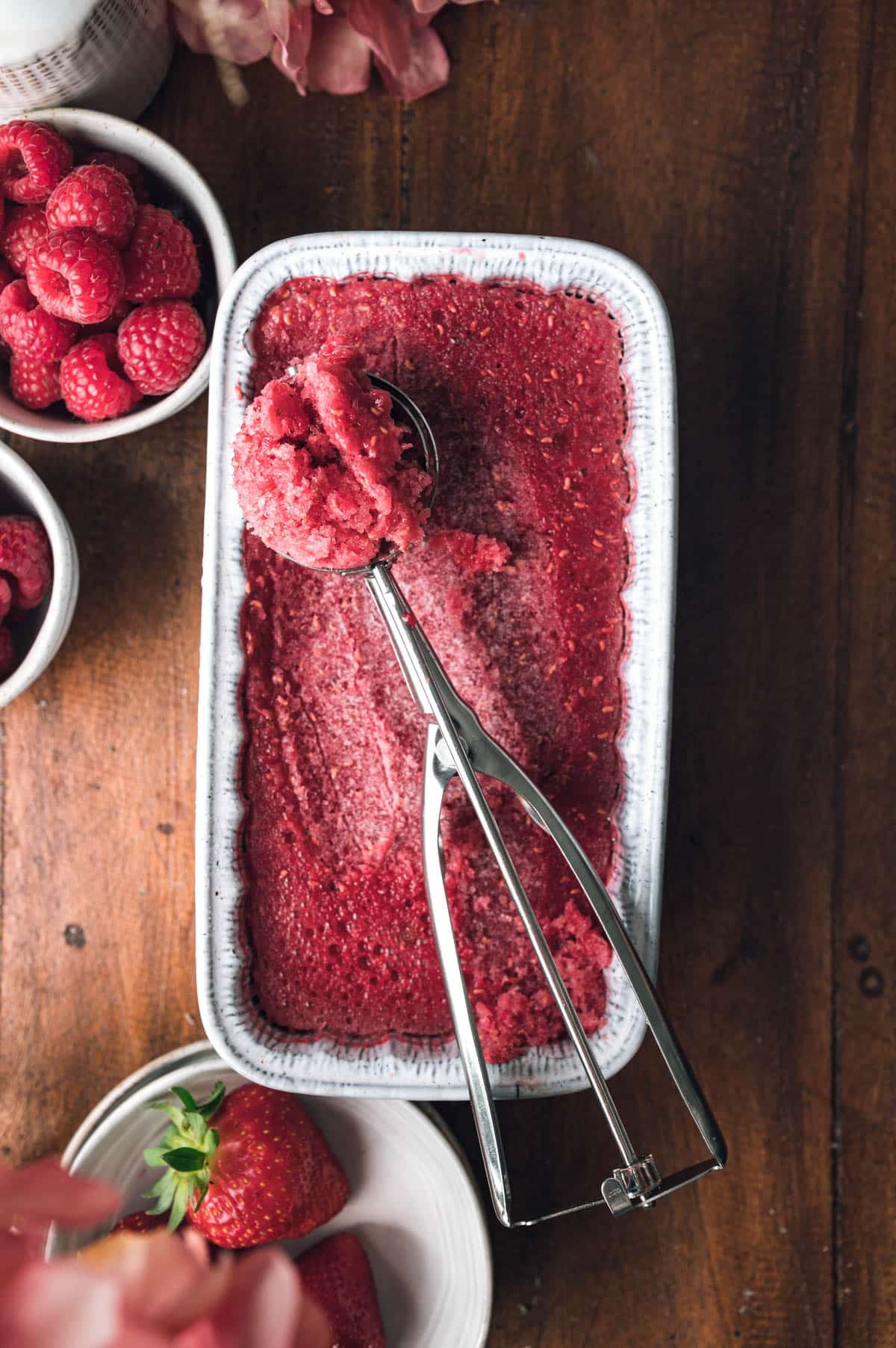 A rectangular dish filled with raspberry sorbet, an ice cream scoop resting on top. Surrounding the dish are bowls holding fresh raspberries and strawberries on a wooden surface.