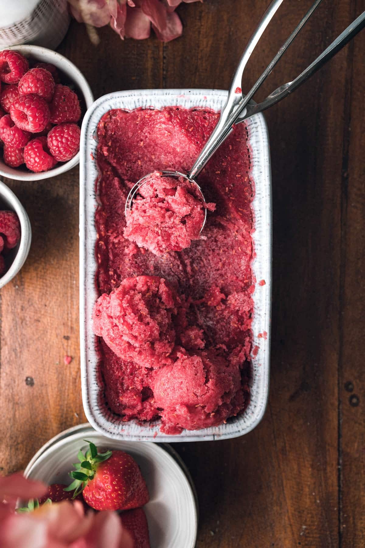 Top view of a rectangular dish filled with raspberry sorbet being scooped. Nearby are bowls containing fresh raspberries and strawberries, all set on a dark wooden surface.