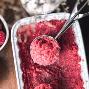 Close-up of a metal scooper in a container of red raspberry sorbet. The sorbet appears freshly scooped, and the container is partially filled. Nearby, there is a bowl of fresh raspberries.