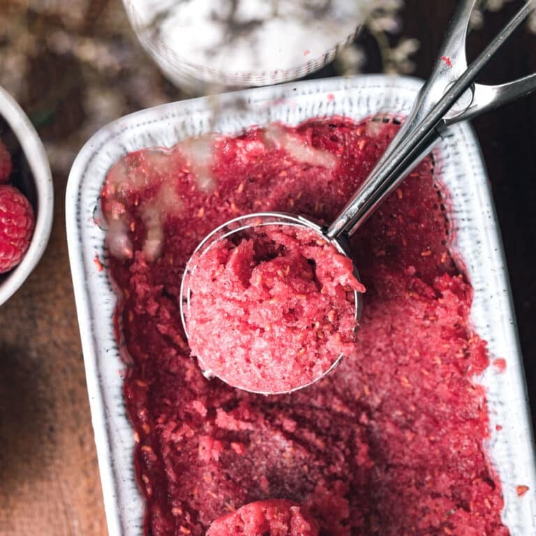 Close-up of a metal scooper in a container of red raspberry sorbet. The sorbet appears freshly scooped, and the container is partially filled. Nearby, there is a bowl of fresh raspberries.