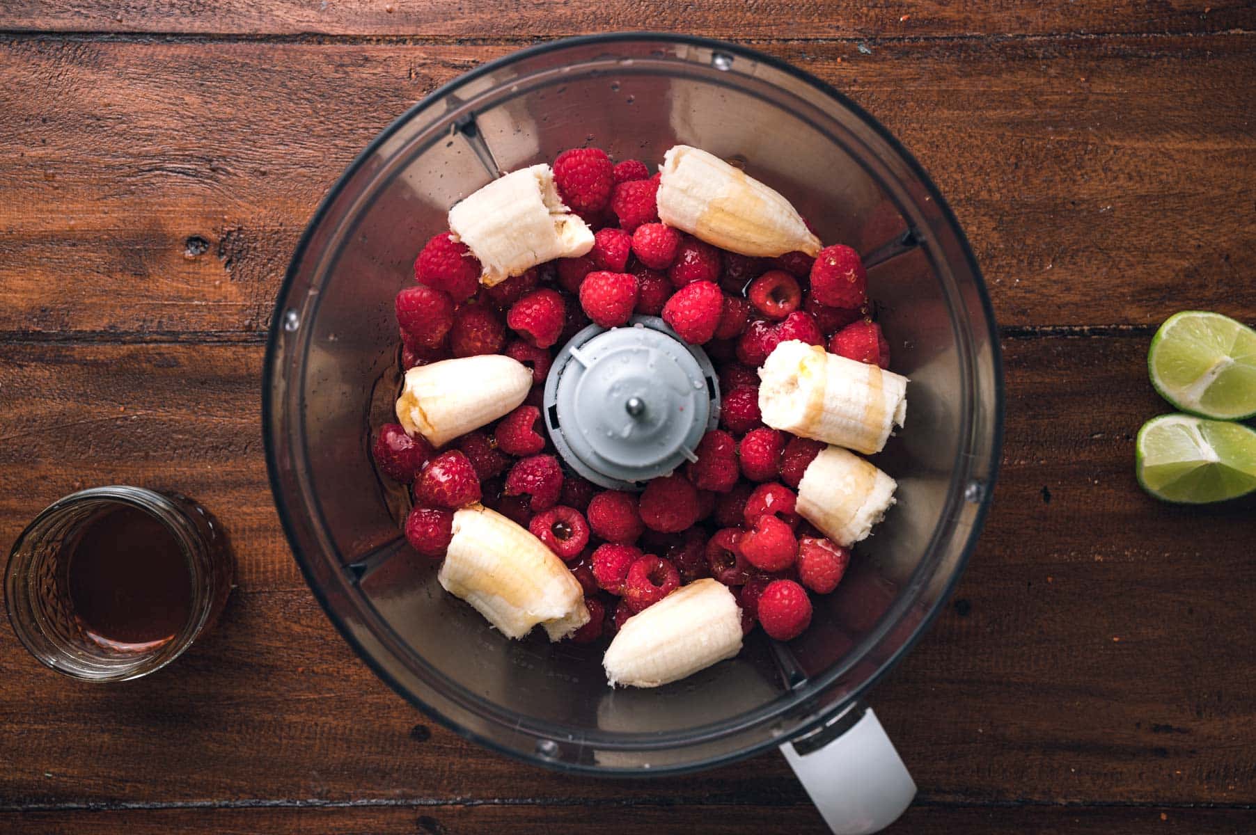 Top view of a food processor with bananas and raspberries; beside the blender, there is a glass of liquid and two lime halves on a wooden surface.
