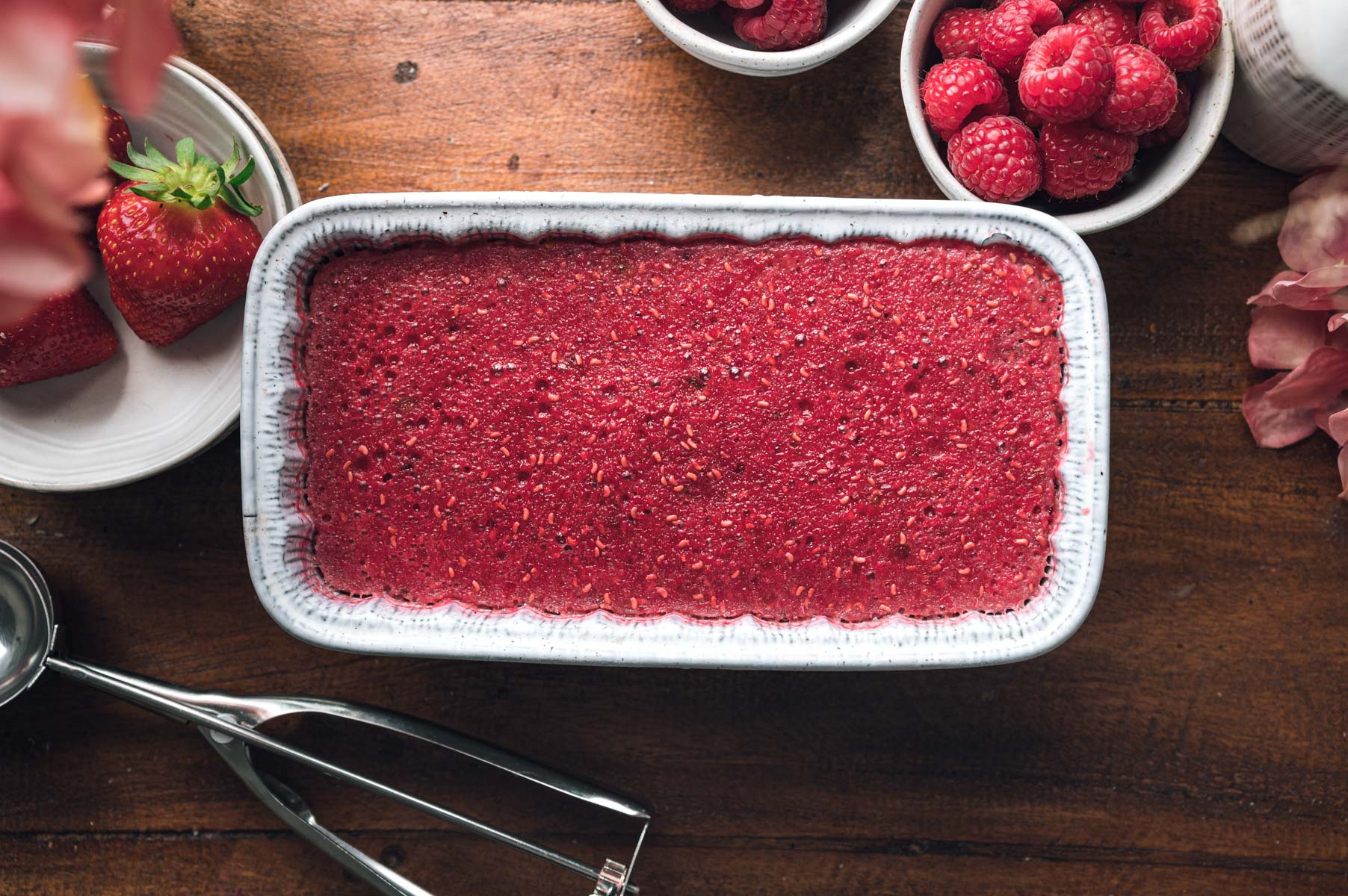 A rectangular dish filled with vibrant red raspberry sorbet is on a wooden table surrounded by small bowls of fresh raspberries, strawberries, and a metal ice cream scoop.