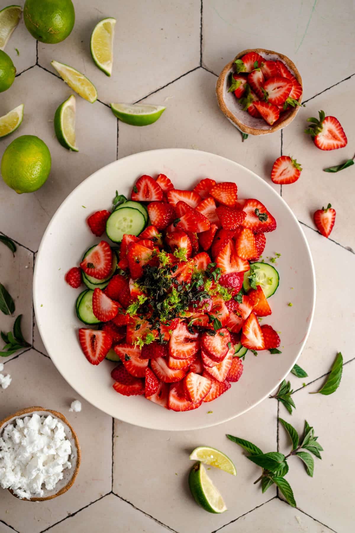 A white bowl filled with a salad of sliced strawberries, cucumber, and mint garnished with lime zest. Surrounding the bowl are lime wedges, mint leaves, and a bowl of crumbled feta cheese.