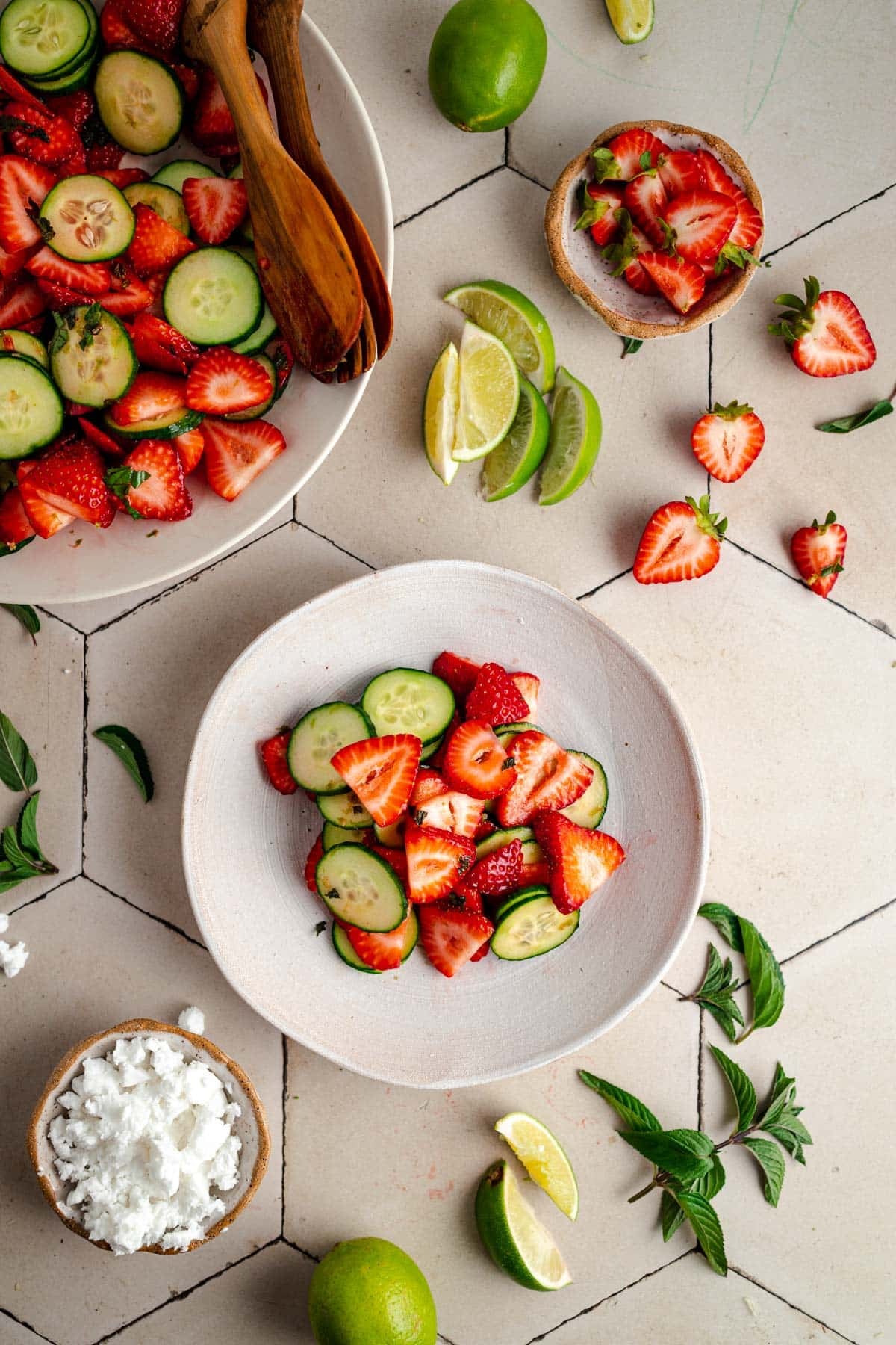 A bowl of cucumber and strawberry salad surrounded by lime wedges, a bowl of crumbled cheese, strawberry halves, sliced cucumber, and fresh herbs on a tiled surface.