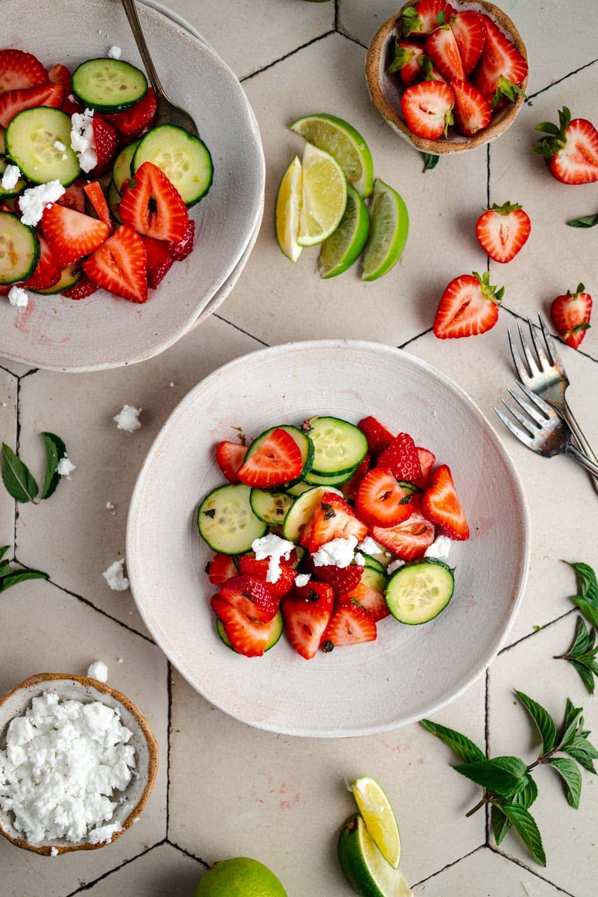 Top-down view of bowls filled with a fresh salad consisting of strawberries, cucumber slices, and a small amount of crumbled cheese, accompanied by lime wedges, herbs, and additional strawberries on the table.