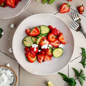 A bowl of salad featuring sliced strawberries, cucumber, and crumbled feta cheese. Fresh mint leaves, lime wedges, and two forks are visible in the background on a tiled surface.