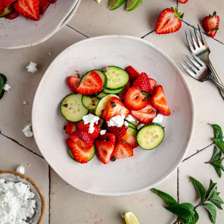 A bowl of salad featuring sliced strawberries, cucumber, and crumbled feta cheese. Fresh mint leaves, lime wedges, and two forks are visible in the background on a tiled surface.