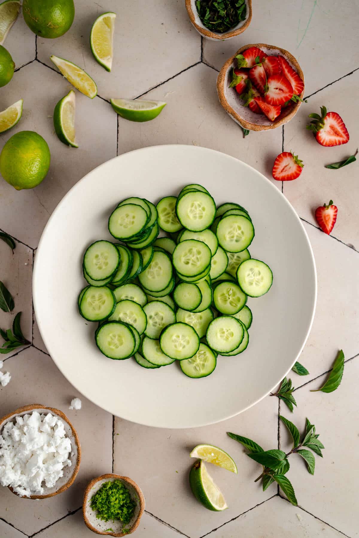 A bowl of sliced cucumbers is centered on a white tiled surface, surrounded by lime wedges, strawberries, mint leaves, and a bowl of feta cheese.