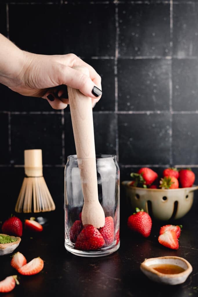 Hand using a wooden muddler to press strawberries in a glass. A bowl of strawberries and other ingredients are visible in the background on a black countertop.