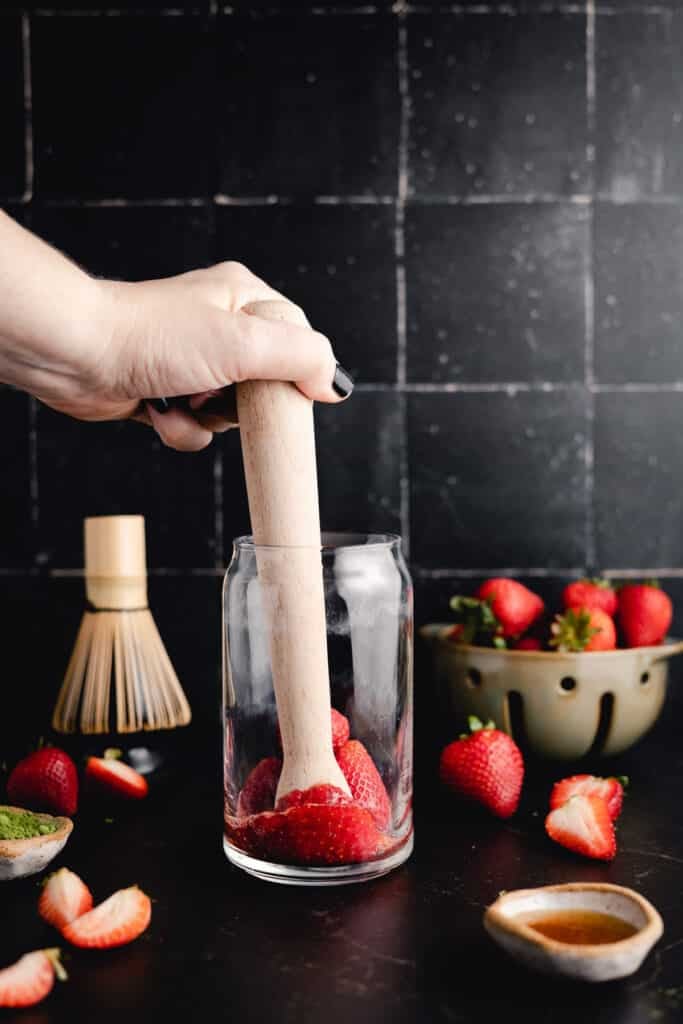 A hand uses a wooden muddler to crush strawberries in a glass. Nearby are whole strawberries in a bowl, a whisk, and a small dish of green powder, all set against a dark tiled background.