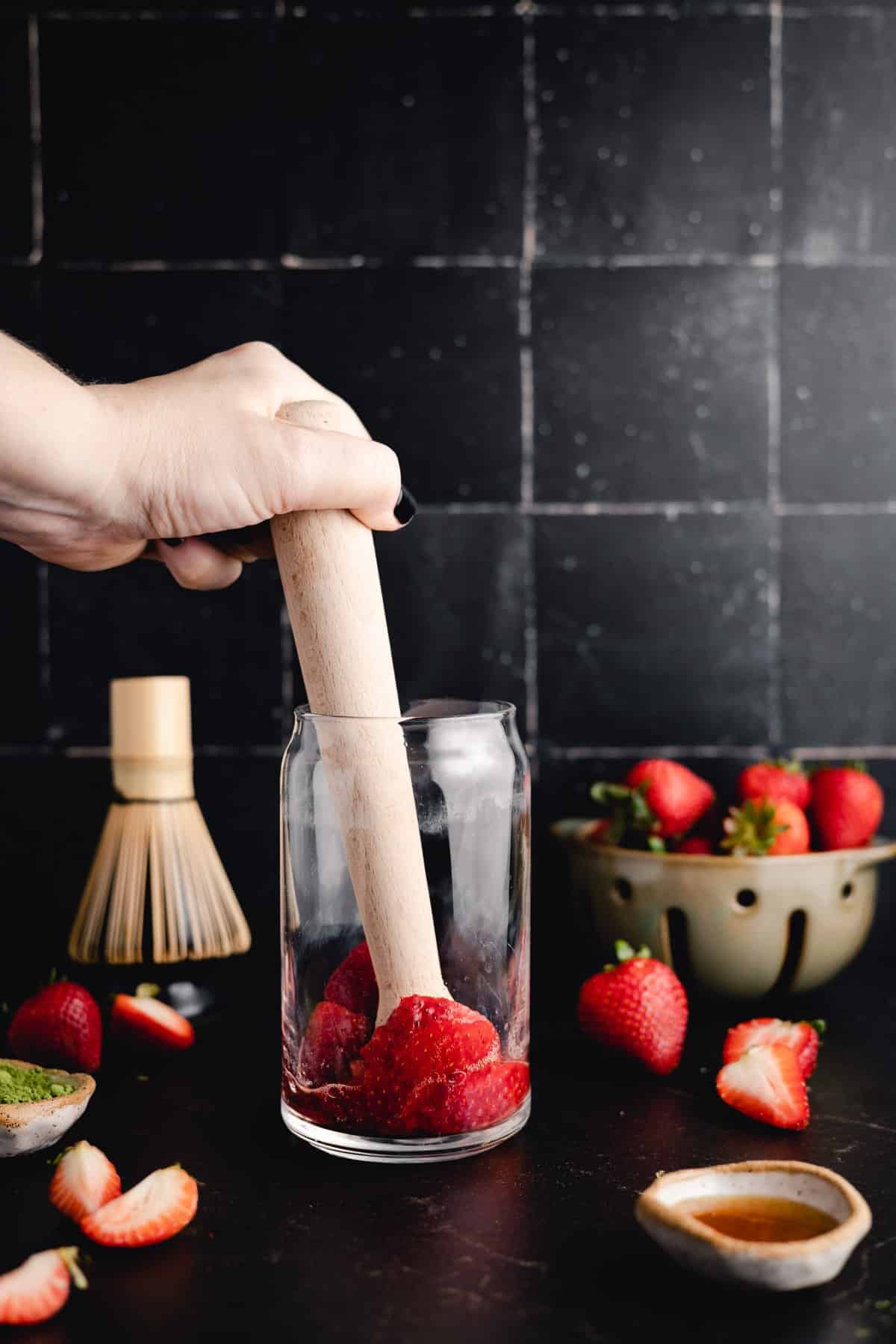A person is using a wooden muddler to crush strawberries in a glass. Nearby are whole strawberries, a bowl of strawberries, a whisk, and a small dish. The background is a tiled wall.