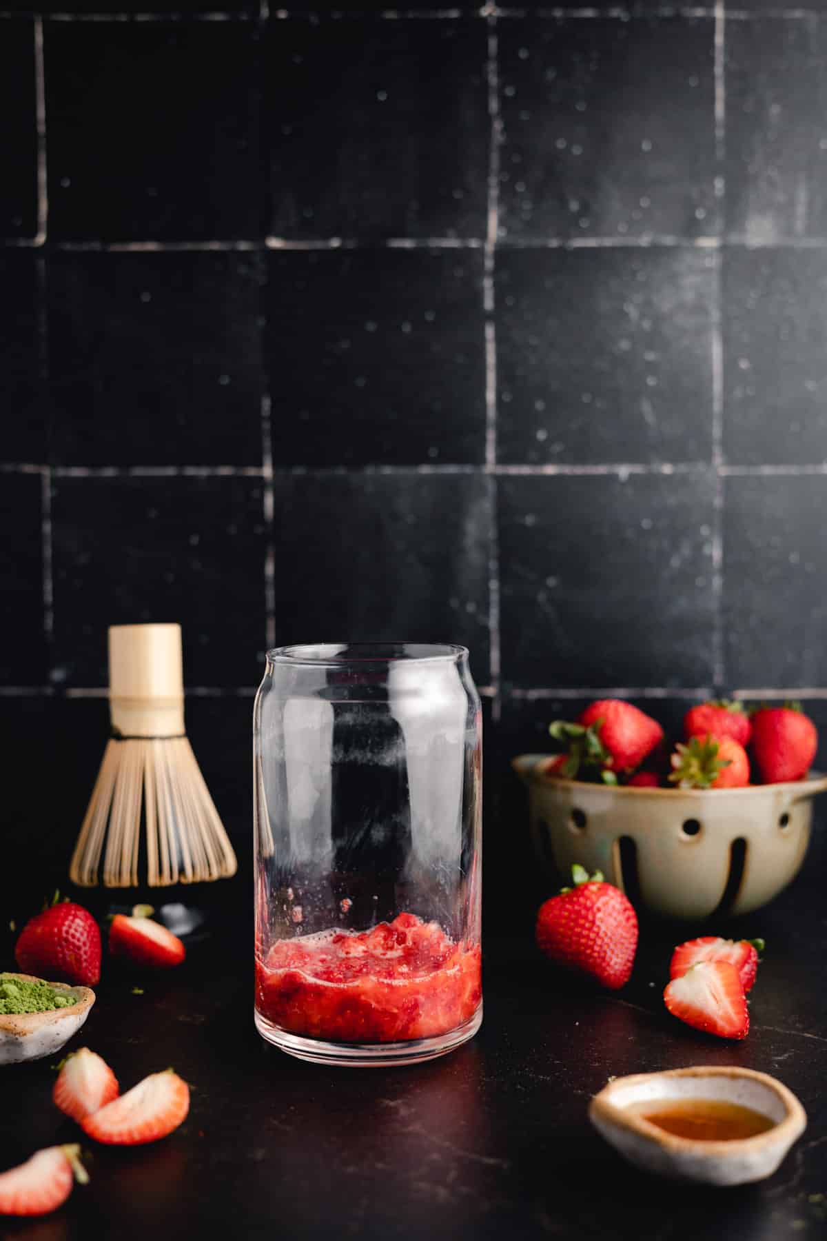 A glass jar with mashed strawberries sits on a black surface. Whole and halved strawberries, small bowls, and a bamboo whisk are arranged around the jar. A bowl of strawberries is in the background.