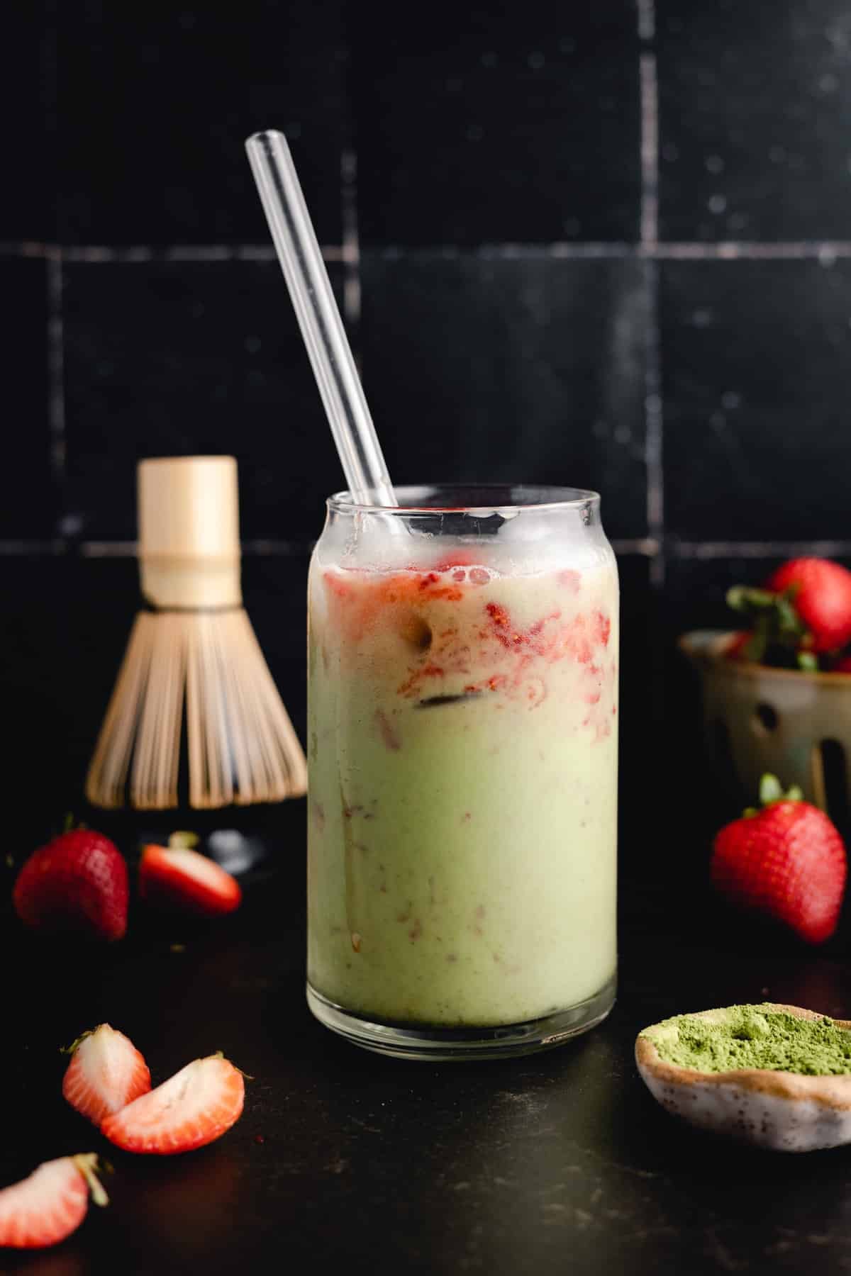 A glass of matcha latte with strawberry puree, topped with ice and a glass straw. Strawberries and a matcha whisk are placed in the background.