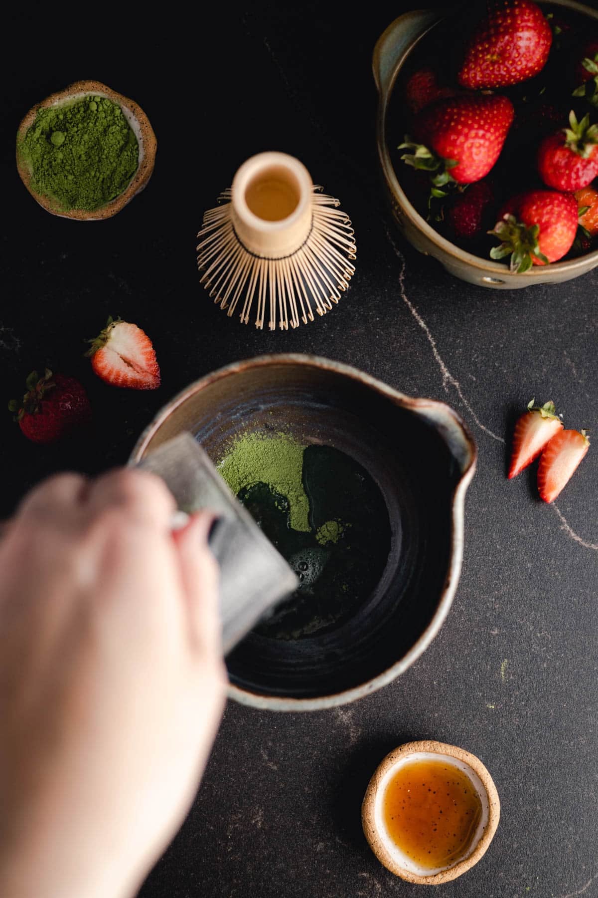 Hand mixing matcha powder in a bowl with a bamboo whisk; strawberries, more matcha powder, and a small bowl of honey are on the table.