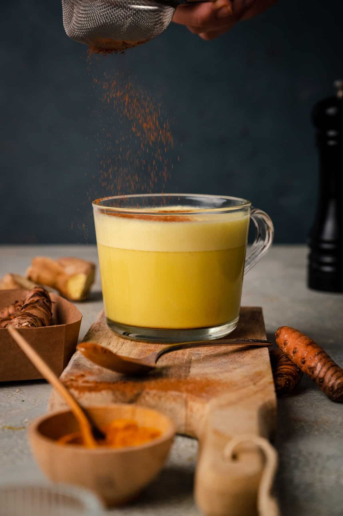 A glass mug of turmeric latte on a wooden board, surrounded by turmeric roots, a bowl of ground turmeric, and a spoon. A hand is sprinkling cinnamon from a sieve onto the latte.