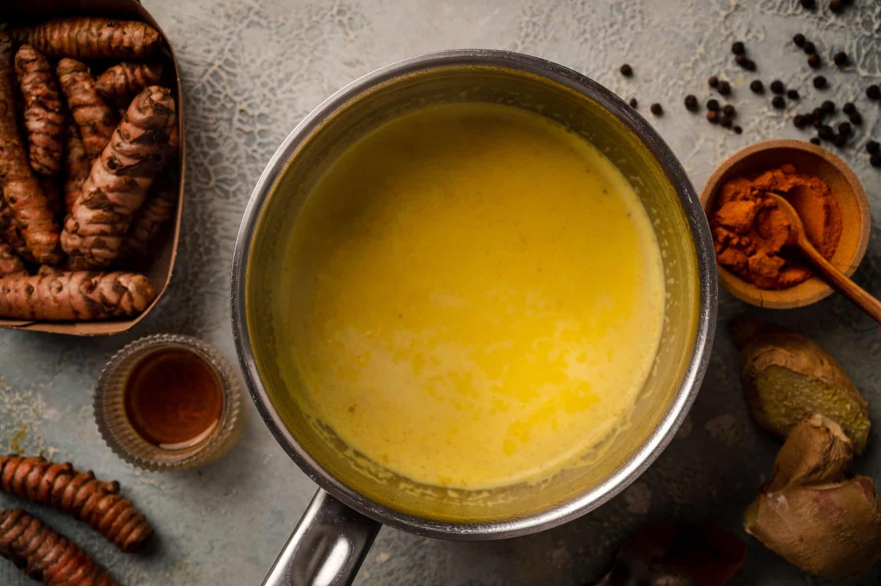 A pot of yellow turmeric milk surrounded by turmeric roots, ground turmeric, ginger, black pepper, and a small cup of honey on a textured surface.