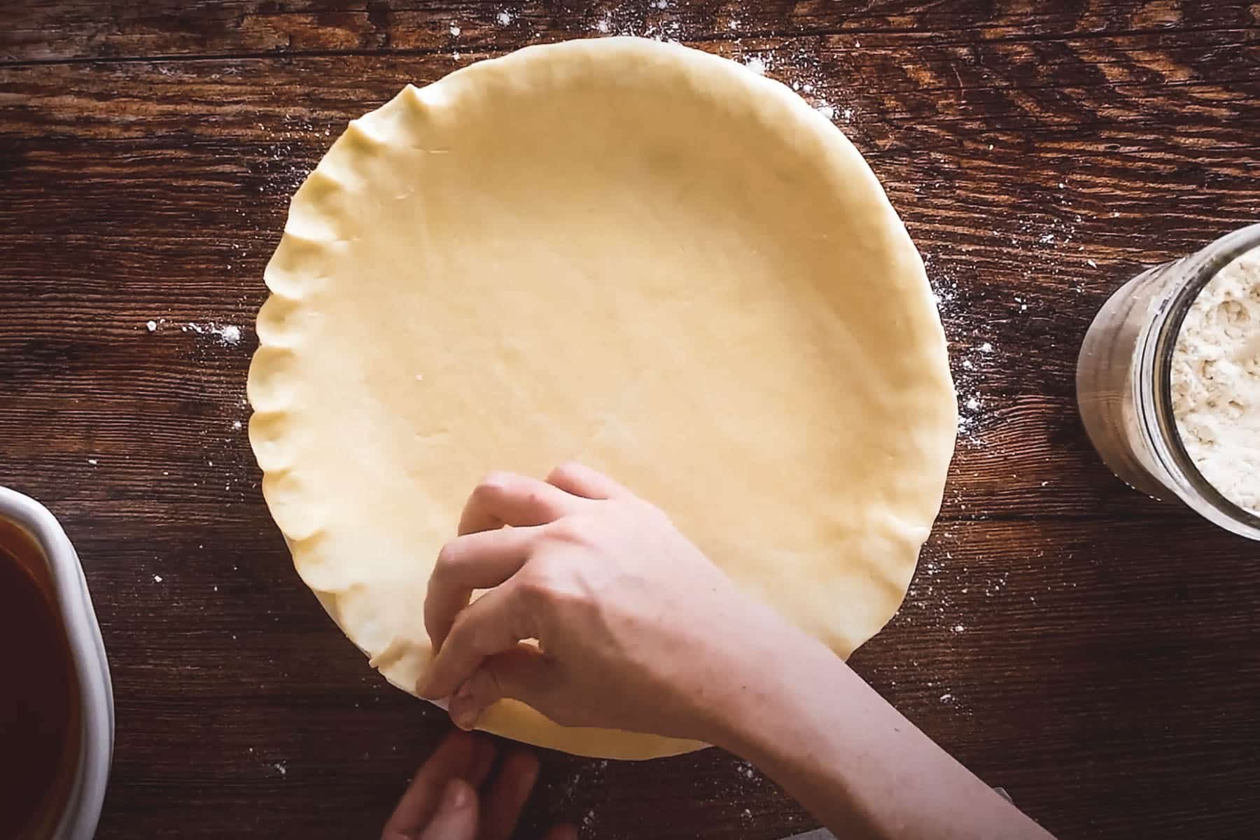 A person crimping the edges of an unbaked pie crust set on a wooden surface.