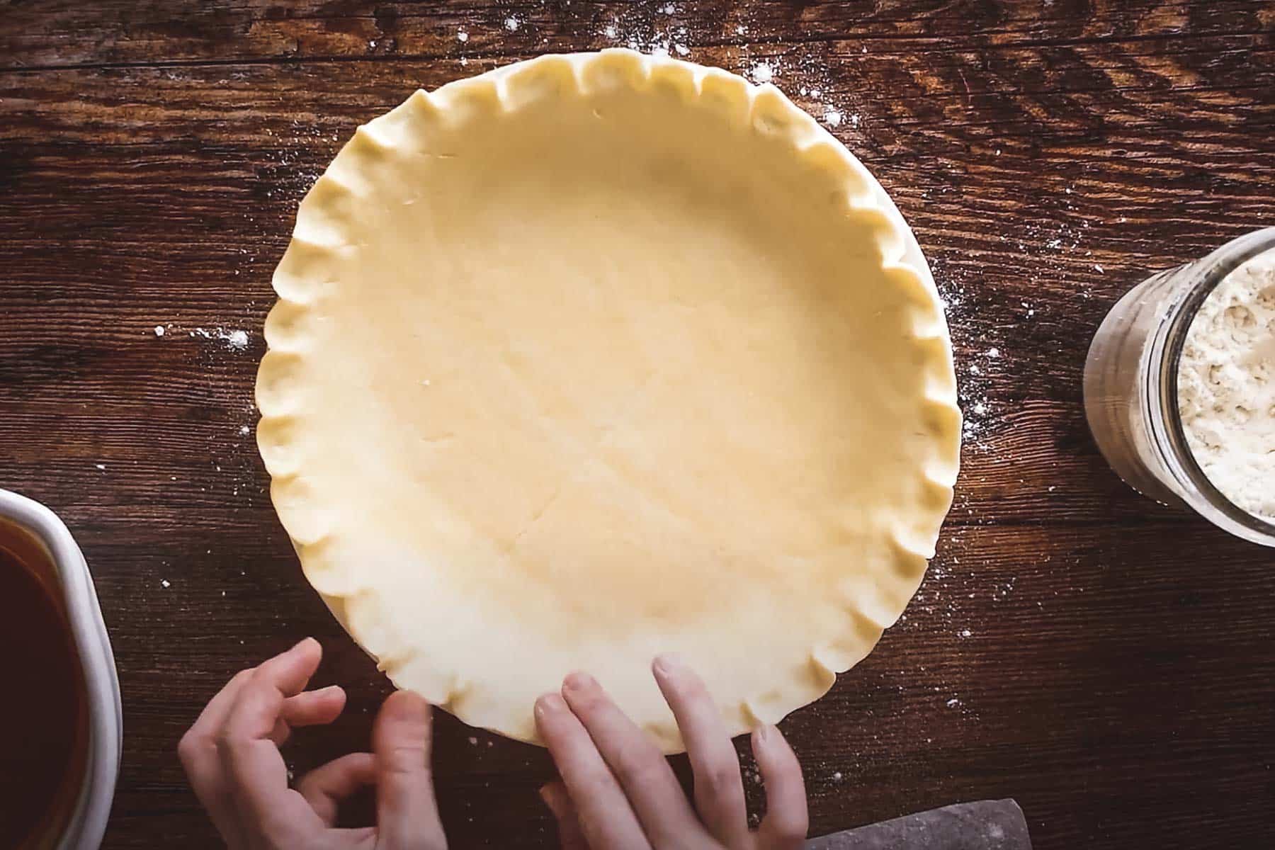 Hands holding an unbaked pie crust on a wooden surface, with a jar of flour and bowl of filling nearby.