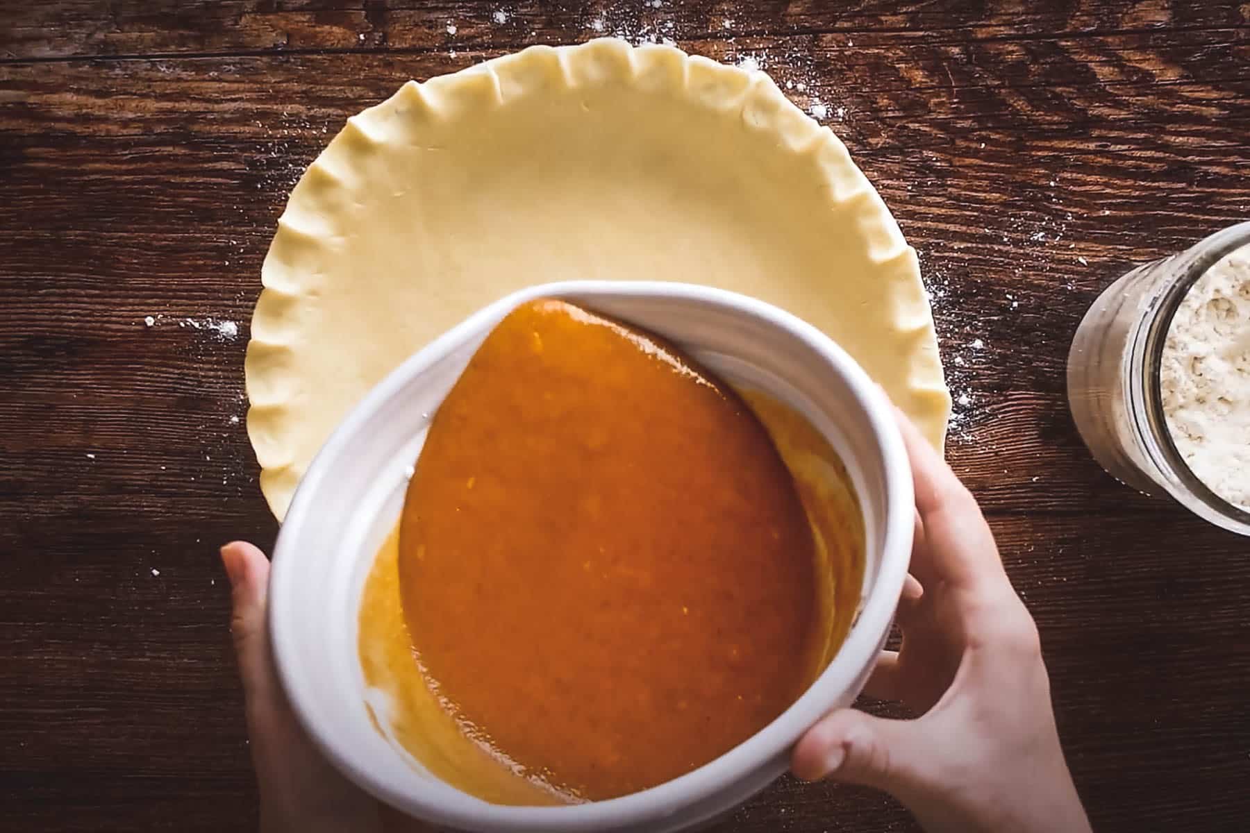 Hands are pouring pumpkin pie filling from a bowl into an unbaked pie crust on a wooden table.