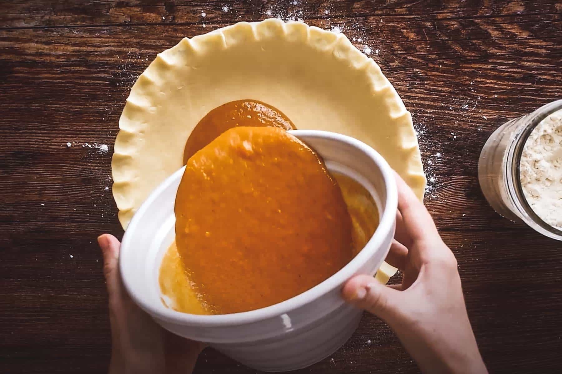 Someone pouring pumpkin filling into an unbaked pie crust on a wooden surface, with flour scattered nearby.