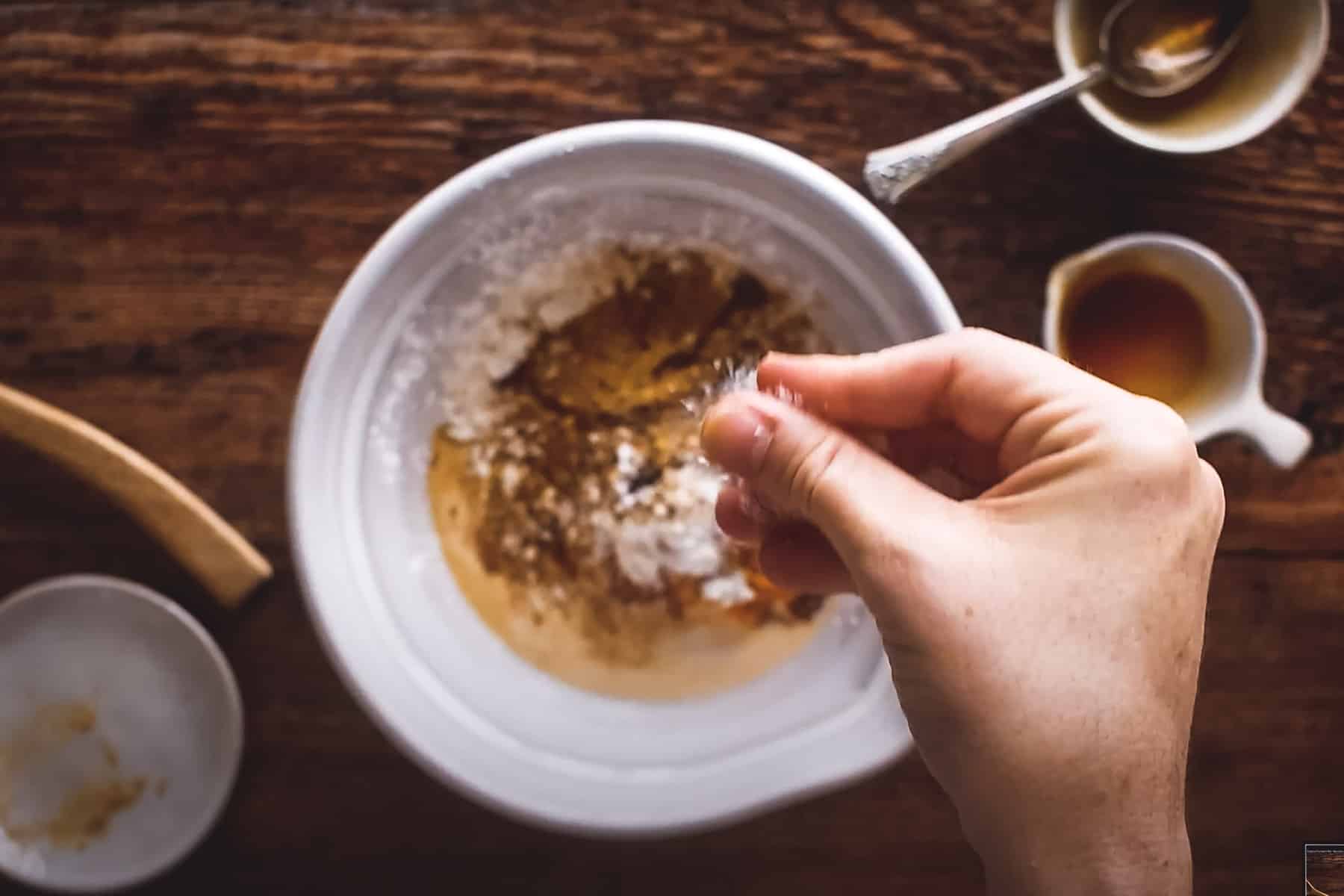 A hand sprinkling salt into a mixing bowl containing flour and pumpkin, etc. with a spoon.